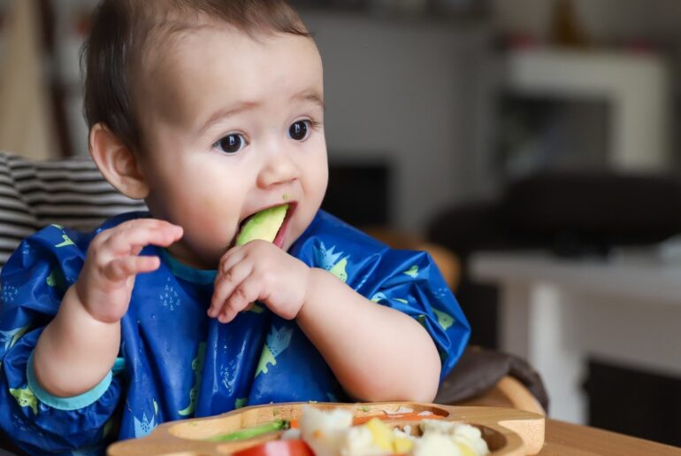 a small baby picks up a piece of cucumber and eats it.