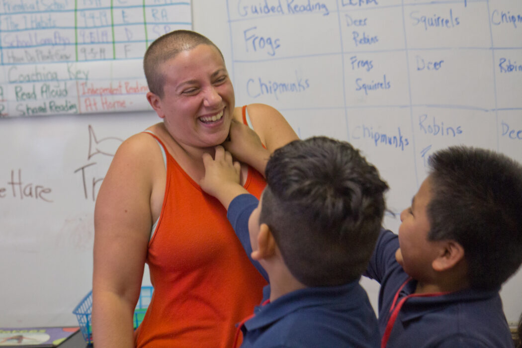 Two young boys in a classroom reach out and tickle their teacher who is laughing.